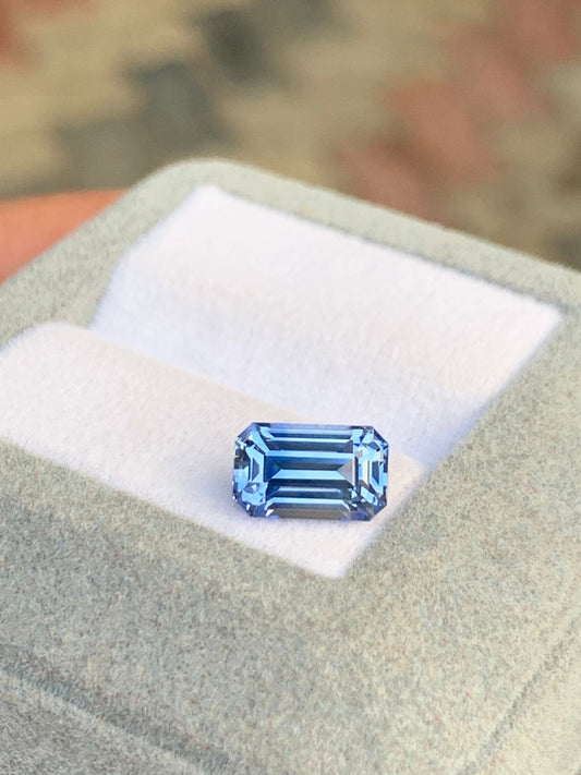 Un superbe saphir bleu taille émeraude de 2,95 carats de Bonnot Paris repose sur un coussin blanc dans une boîte à bijoux gris clair avec un fond flou, mettant en valeur sa beauté naturelle non chauffée du Sri Lanka.