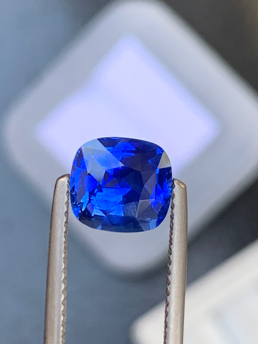 Gros plan sur le Saphir bleu • 3,00 ct de [NOUVEAU] Bonnot Paris : un saphir bleu profond et éclatant, saisi dans une pince de joaillier. La pierre facettée reflète la lumière, révélant sa clarté et sa couleur intense sur un fond flou, soulignant ainsi sa beauté intemporelle.