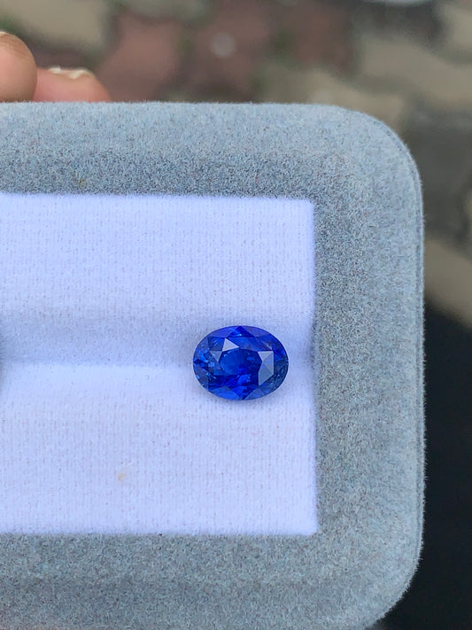 Une pierre précieuse ovale bleue vibrante, le Saphir bleu de 3,55 ct de Bonnot Paris, est présentée sur une surface coussinée blanche et grise douce, faisant peut-être partie d'une boîte à bijoux ou d'une vitrine.