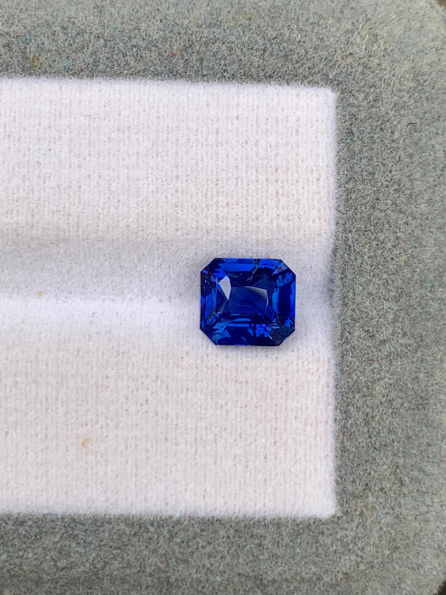 Un saphir bleu taille émeraude d'un bleu profond de 2,53 carats, provenant du Sri Lanka, signé [NOUVEAU] Bonnot Paris, repose sur un fond de tissu blanc et vert, mettant en valeur ses facettes brillantes et sa couleur intense. Cette pierre précieuse non chauffée séduit par son charme naturel.