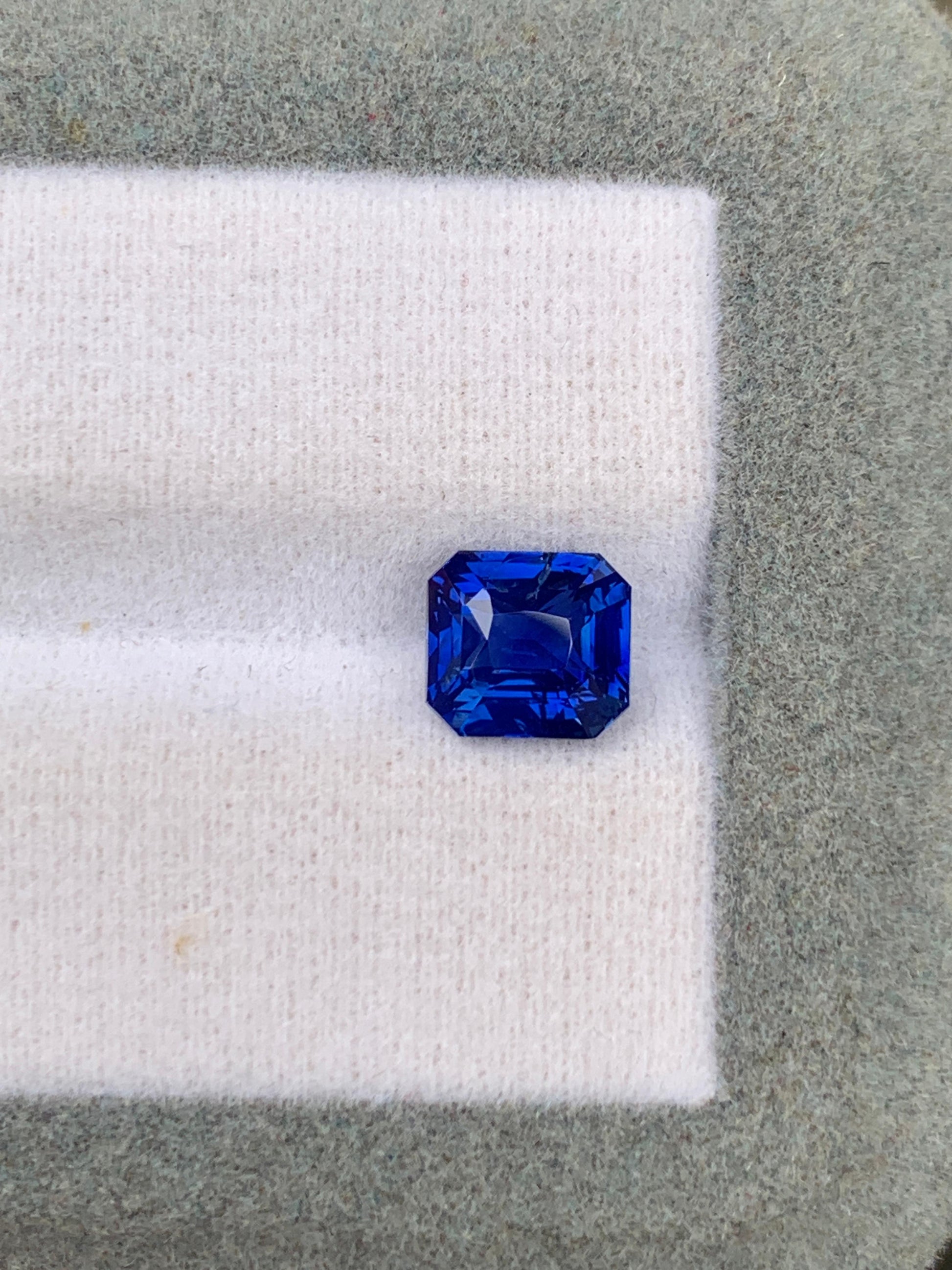 Un saphir bleu taille émeraude d'un bleu profond de 2,53 carats, provenant du Sri Lanka, signé [NOUVEAU] Bonnot Paris, repose sur un fond de tissu blanc et vert, mettant en valeur ses facettes brillantes et sa couleur intense. Cette pierre précieuse non chauffée séduit par son charme naturel.