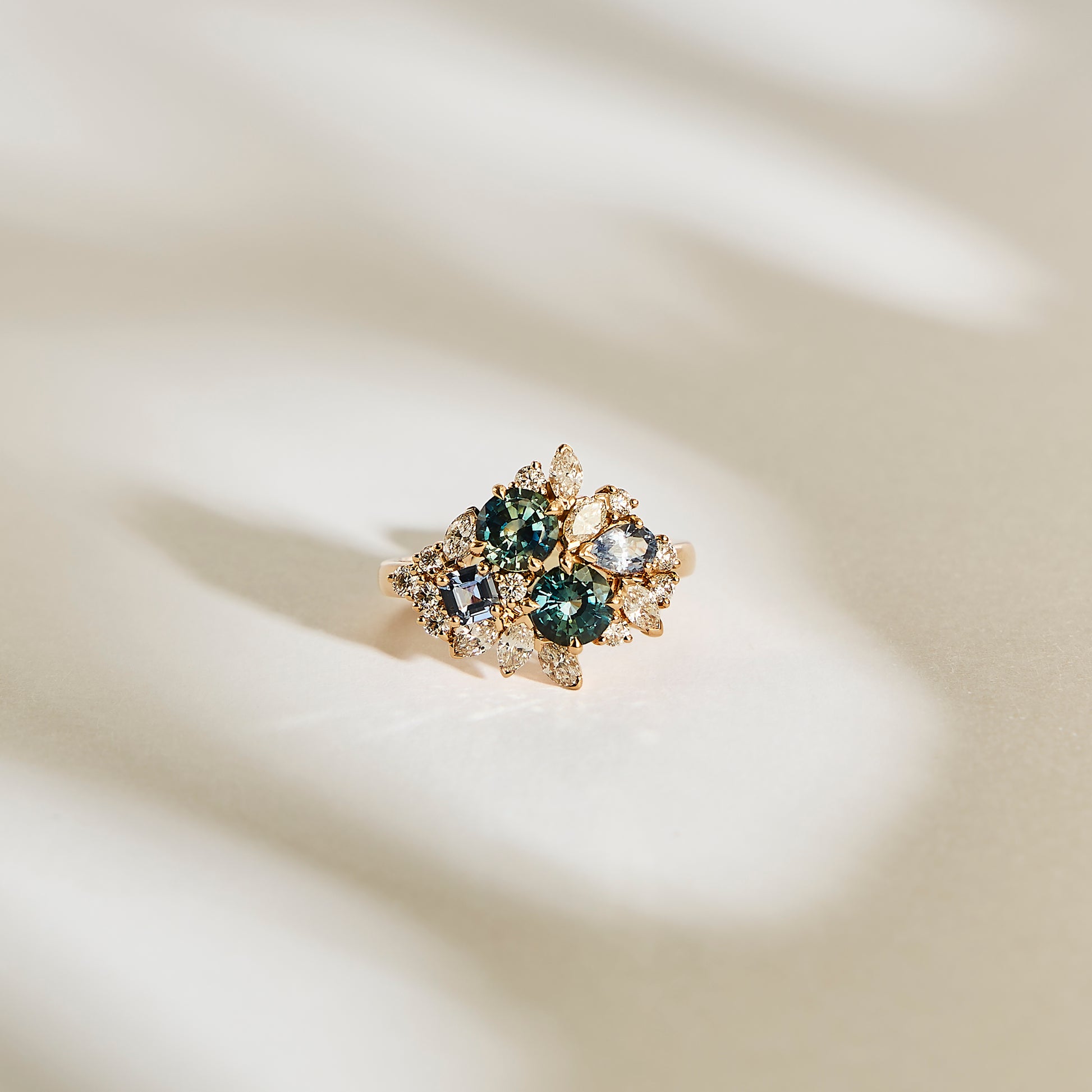 La bague "Teal Bloom - Saphir" de Bonnot Paris est une bague en or ornée de saphirs Teal et de pierres précieuses claires dans un design floral, joliment sertie sur un fond doux et clair avec des ombres douces.