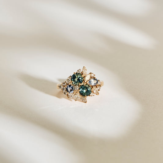 La bague "Teal Bloom - Saphir" de Bonnot Paris est une bague en or ornée de saphirs Teal et de pierres précieuses claires dans un design floral, joliment sertie sur un fond doux et clair avec des ombres douces.