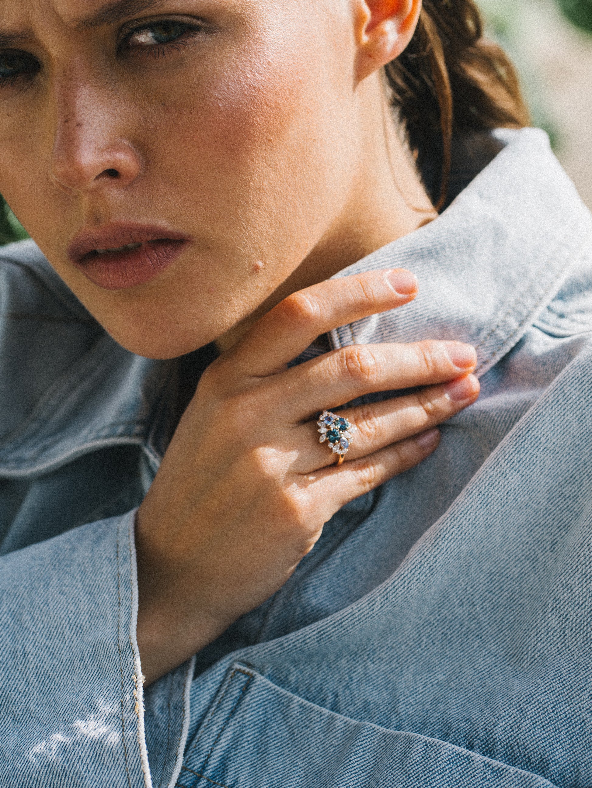 Vêtue d'une veste en jean légère, quelqu'un touche sa poitrine pour révéler la bague Bonnot Paris "Teal Bloom - Saphir", comportant une pierre précieuse verte frappante entourée de diamants scintillants sur un fond légèrement flou.