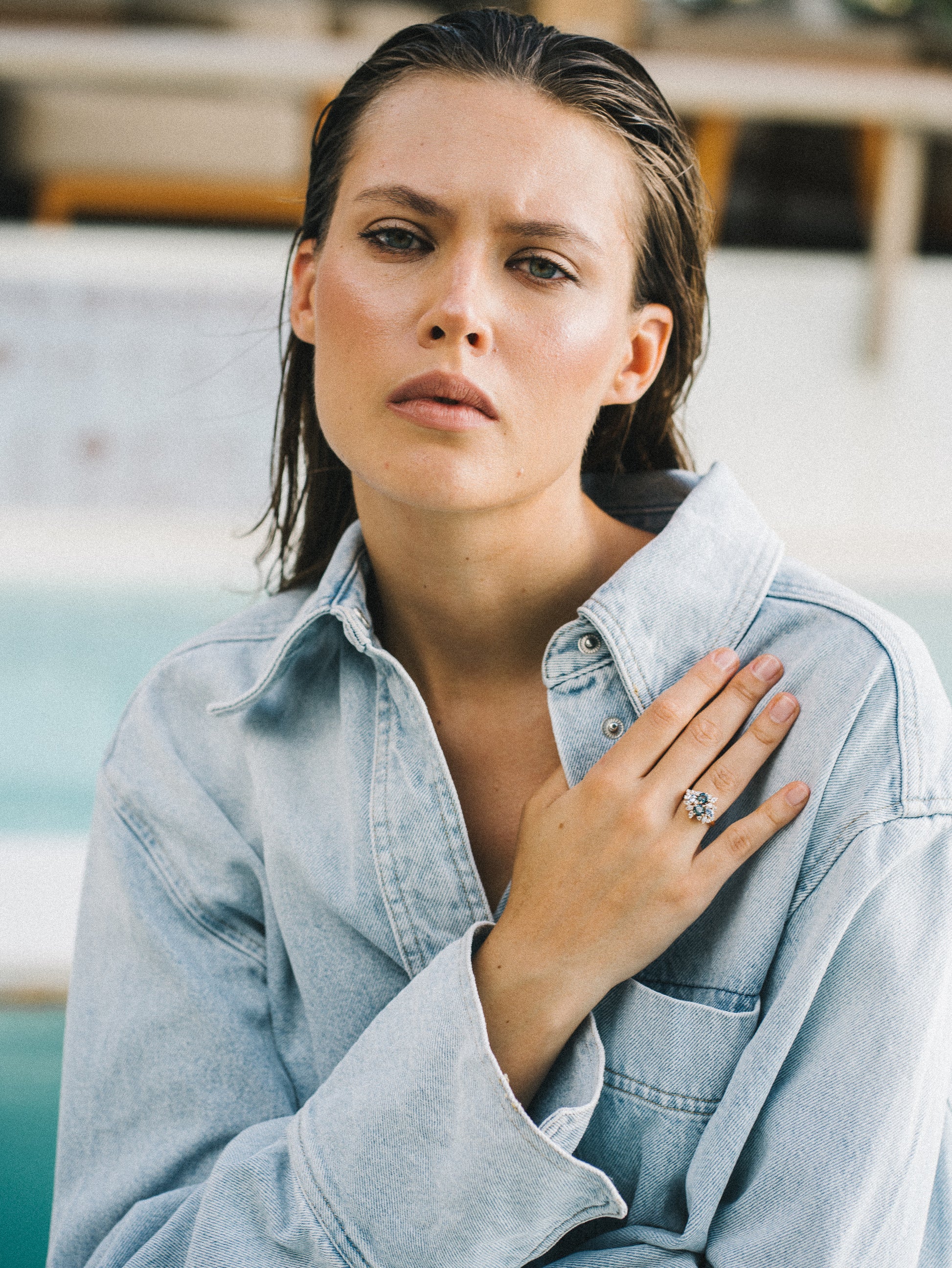Une personne, les cheveux mouillés, vêtue d'une veste en jean légère, pose au bord d'une piscine, regardant droit vers l'objectif. Une main posée sur sa poitrine met en valeur la bague Teal Bloom - Saphir de Bonnot Paris. L'arrière-plan est légèrement flou.