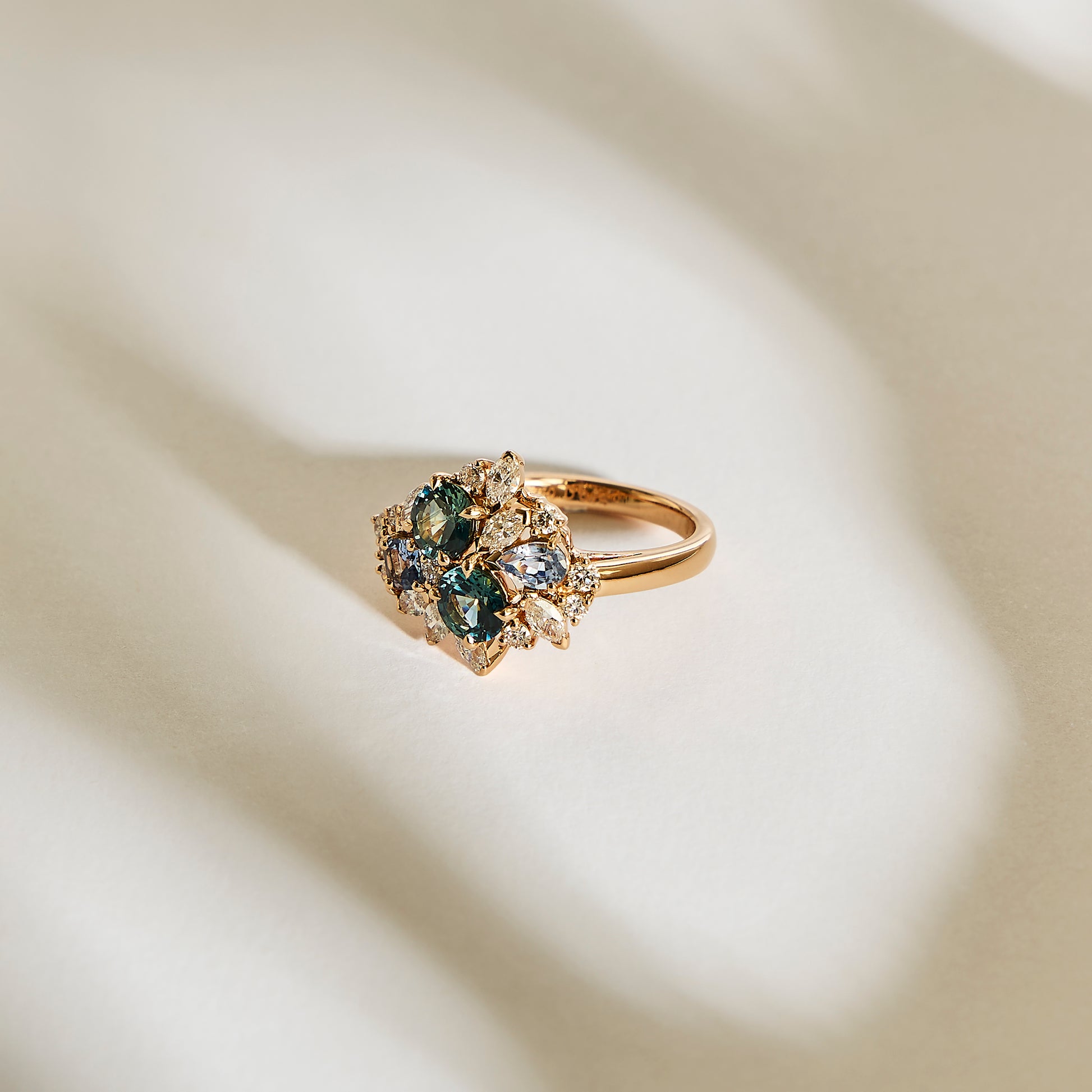 La bague en or Teal Bloom - Saphir de Bonnot Paris, ornée de saphirs bleu sarcelle et de diamants blancs, repose sur une surface crème doucement éclairée avec un groupe complexe de pierres précieuses projetant des ombres douces.