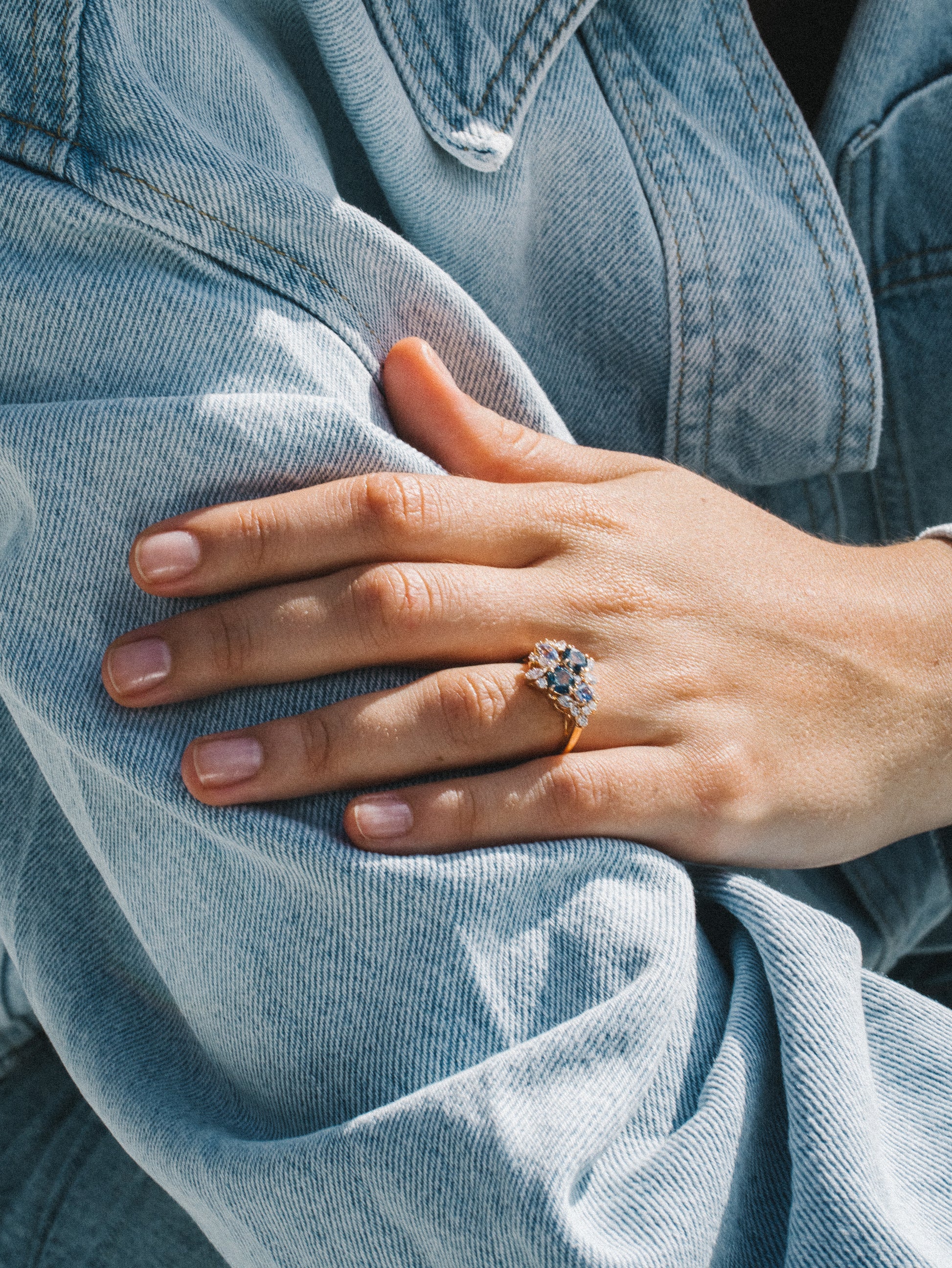 Une personne portant une veste en jean légère pose sa main sur son bras, mettant en valeur l'élégante bague Bonnot Paris Teal Bloom - Saphir avec une grosse pierre précieuse murmurant des diamants et des saphirs.