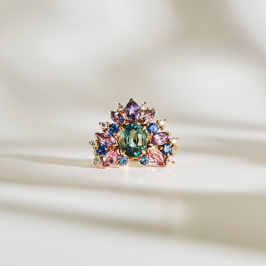 L'Indigo Bloom - Saphir de Bonnot Paris, façonné dans le style de la haute joaillerie, met en valeur une pierre précieuse verte centrale entourée de pierres violettes, bleues et roses serties d'or. Des diamants scintillants rehaussent son élégance tandis qu'il repose sur une surface blanche, projetant des ombres délicates qui accentuent son design complexe.