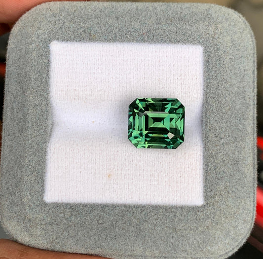 L'exquis Saphir vert • 10,55ct de [NOUVEAU] Bonnot Paris brille dans une boîte grise carrée, sa taille émeraude précise et sa teinte bleu sarcelle profonde mises en valeur sur un tissu blanc moelleux, créant une pièce maîtresse captivante.