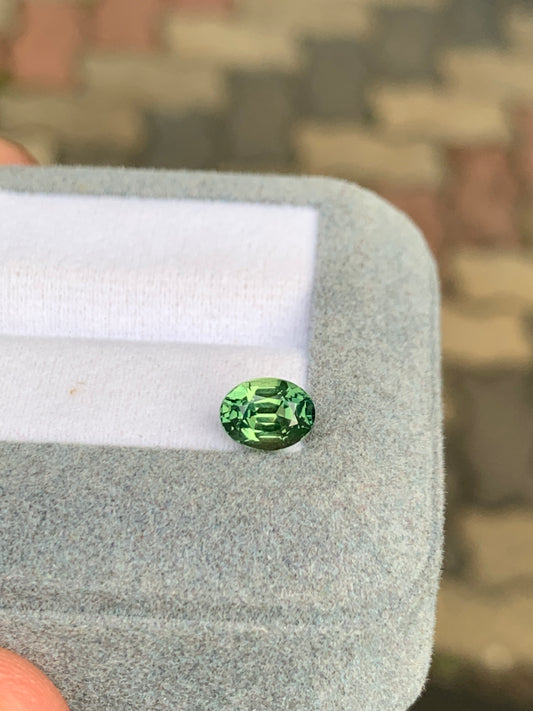 Un Saphir vert de 1,55 carat de Bonnot Paris repose élégamment sur le bord d'un coffret à bijoux gris clair sur un fond flou au motif de pavés, évoquant le riche paysage de Madagascar.