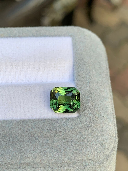 Un Saphir Vert taille émeraude rectangulaire de 3,15 carats de Bonnot Paris repose sur un présentoir coussiné gris clair, avec des motifs verts et des stries plus foncées créant une superbe tapisserie visuelle sur sa surface.