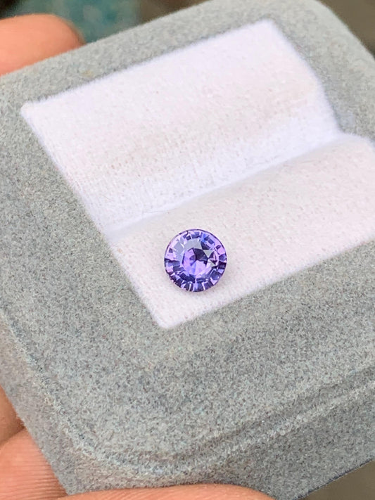 Le Saphir violet • 1,03ct de [NOUVEAU] Bonnot Paris, une pierre précieuse lavande à facettes rondes apparentée à la variété sri-lankaise, est élégamment présenté sur une surface coussinée blanche à l'intérieur d'une boîte en velours gris, délicatement tenue entre les doigts.