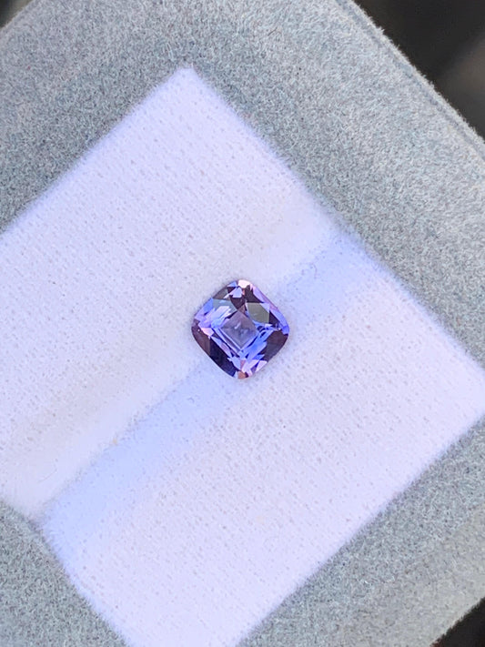 Un saphir violet taille coussin de 1,08 ct de chez Bonnot Paris est posé au centre d'un écrin gris clair doublé de tissu blanc. La pierre scintille sous la lumière, accentuant sa couleur vive et ses facettes.