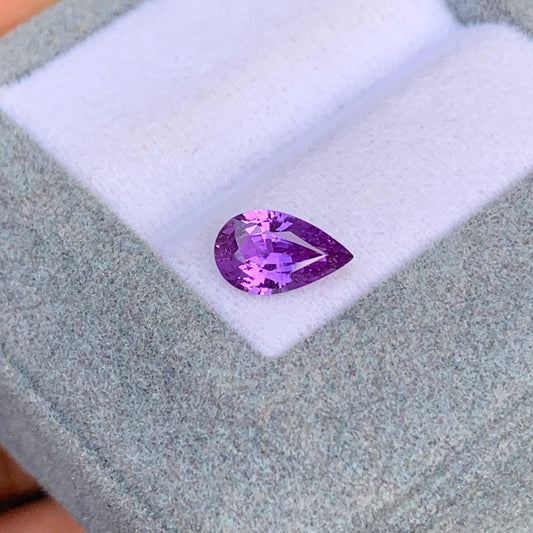 Une pierre précieuse violette Saphir (1,18 ct) de [NOUVEAU] Bonnot Paris repose sur un tissu gris clair dans une boîte à bijoux ouverte, sa couleur vibrante et ses facettes brillant de clarté et de qualité rappelant les célèbres pierres précieuses du Sri Lanka.