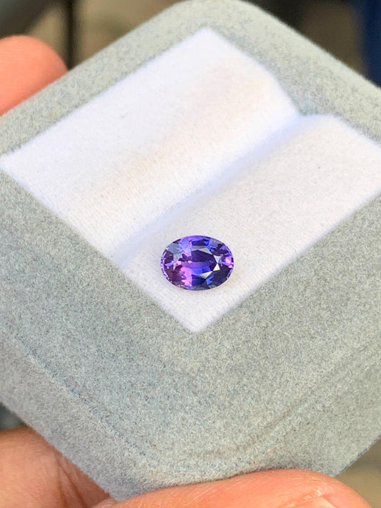 Une main ouvre un écrin ouvert révélant une pierre précieuse violette taille ovale de 1,26 carat de la maison Bonnot Paris. Le saphir violet repose sur un coussin blanc dans un écrin gris clair à la texture douce, reflétant la lumière pour mettre en valeur ses facettes, à l'image de la haute joaillerie sri-lankaise.