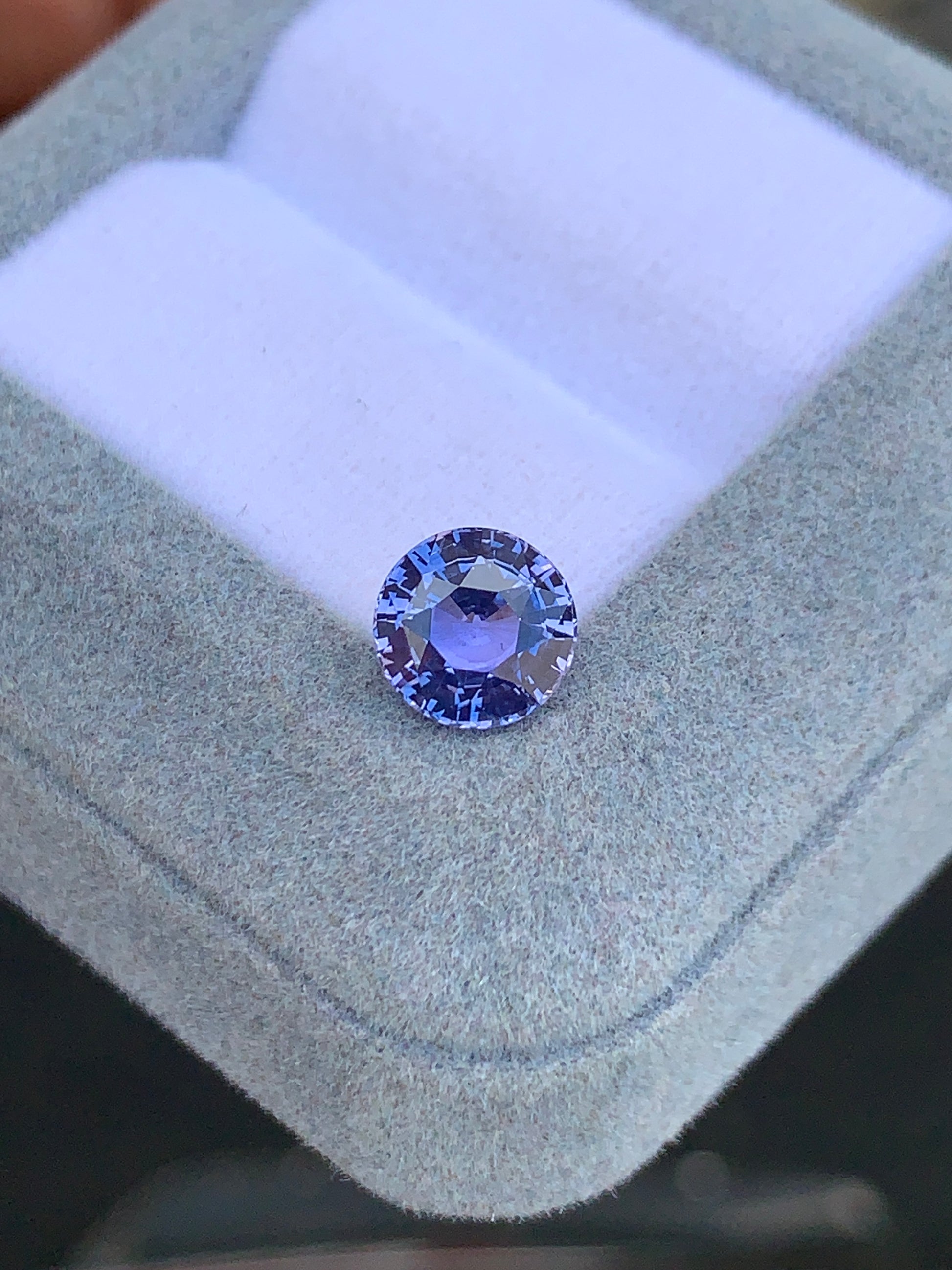 Le Saphir violet rond facetté • 2,45ct de [NOUVEAU] Bonnot Paris scintille sur un coussin blanc à l'intérieur d'une boîte gris clair, mettant en valeur sa clarté et sa taille complexe parmi d'autres pierres précieuses.