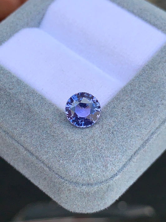 Le Saphir violet rond facetté • 2,45ct de [NOUVEAU] Bonnot Paris scintille sur un coussin blanc à l'intérieur d'une boîte gris clair, mettant en valeur sa clarté et sa taille complexe parmi d'autres pierres précieuses.