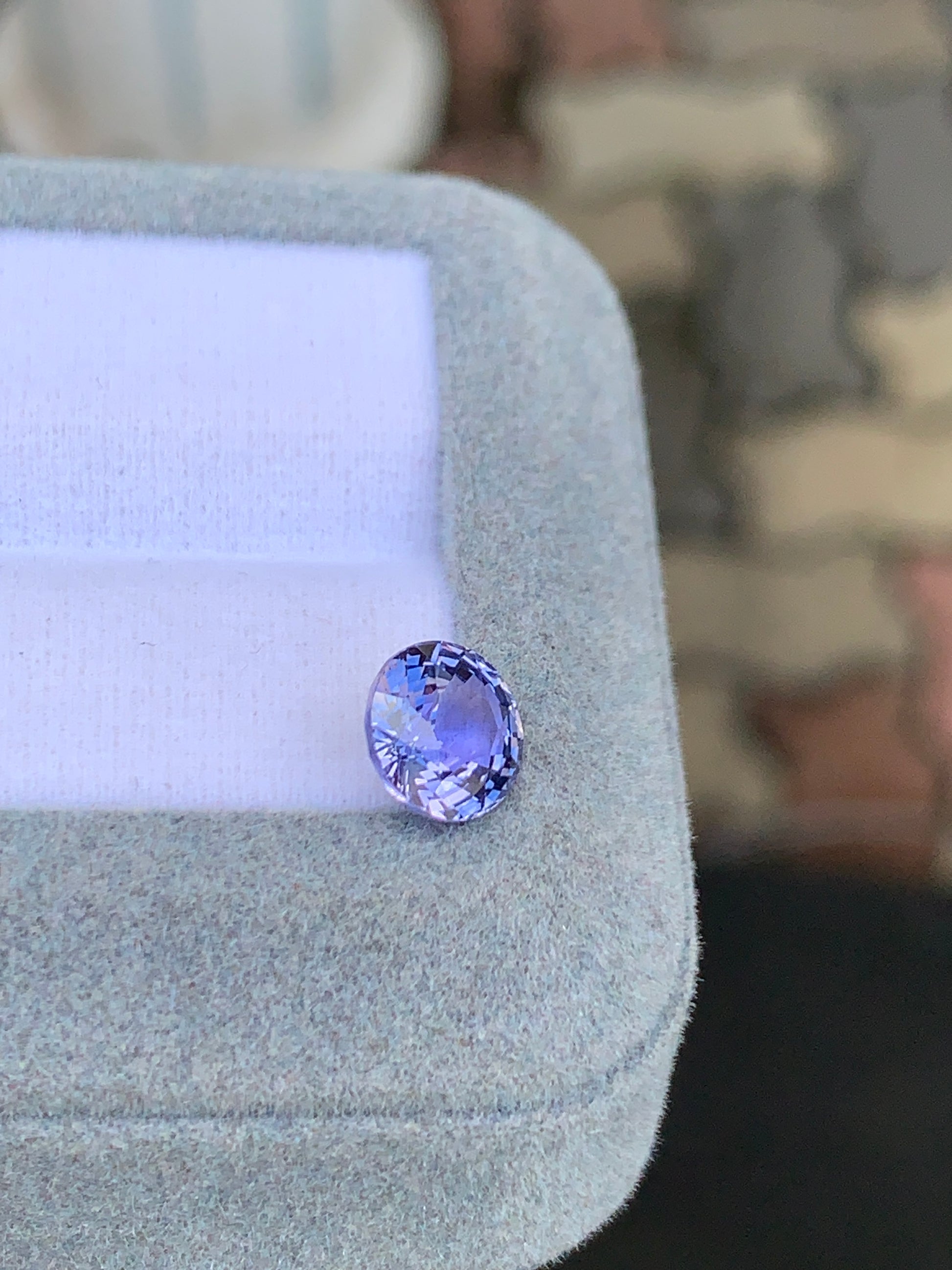 La pierre précieuse violette Saphir de 2,45 ct de Bonnot Paris, une beauté lavande de taille ovale, est présentée sur un coussin gris clair avec un fond à motif brique flou, ses facettes scintillant à la lumière.