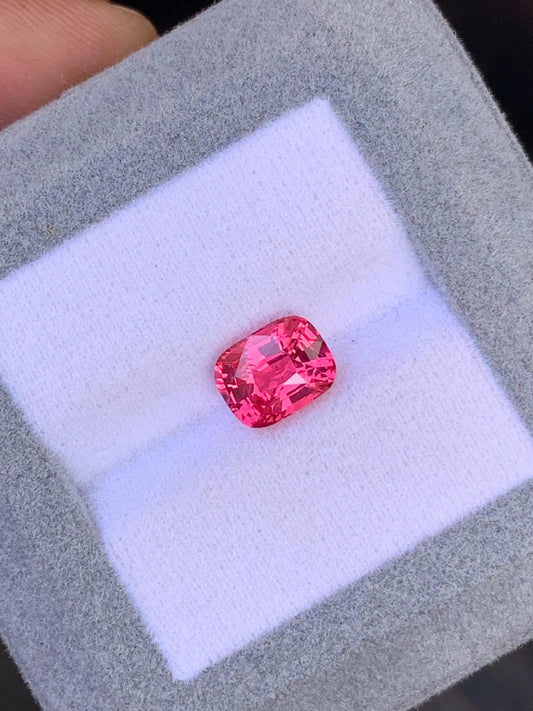 Un spinelle Mahenge taille coussin éclatant, provenant des célèbres mines de Mahenge (2,53 carats), scintille sur un tissu blanc, dans un écrin gris signé Bonnot Paris. Ses facettes, bien visibles, rehaussent son éclat et sa couleur, mettant en valeur sa beauté exquise.