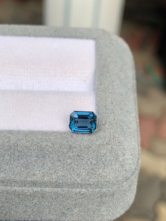 Un spinelle bleu taille émeraude réfléchissant de 1,03 carat de Bonnot Paris repose sur un coussin gris. Sa surface polie contraste avec la texture douce du coussin sur un fond légèrement flou.