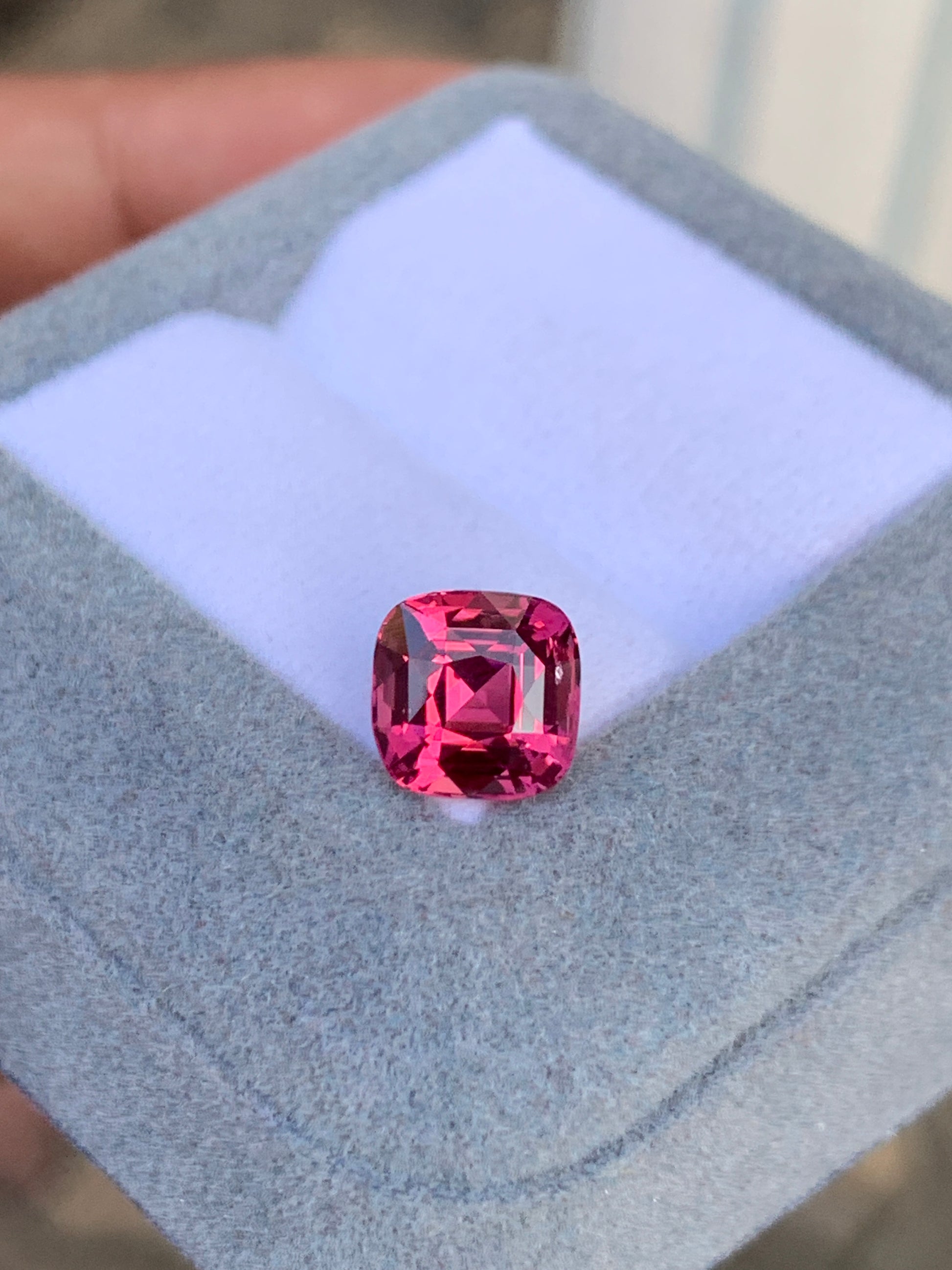 Une pierre précieuse spinelle rose Mahenge de Bonnot Paris, d'un rose éclatant de 3,02 carats, est présentée dans un écrin gris ouvert doublé de blanc. Cette pierre taille coussin révèle toute sa brillance en captant la lumière grâce à ses multiples facettes.
