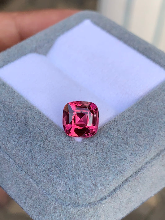 Une pierre précieuse rose Spinelle vibrante de 3,02 ct de [NOUVEAU] Bonnot Paris, taille coussin, repose sur une plate-forme coussinée blanche à l'intérieur d'une élégante boîte à bijoux grise, élégamment tenue à la main.