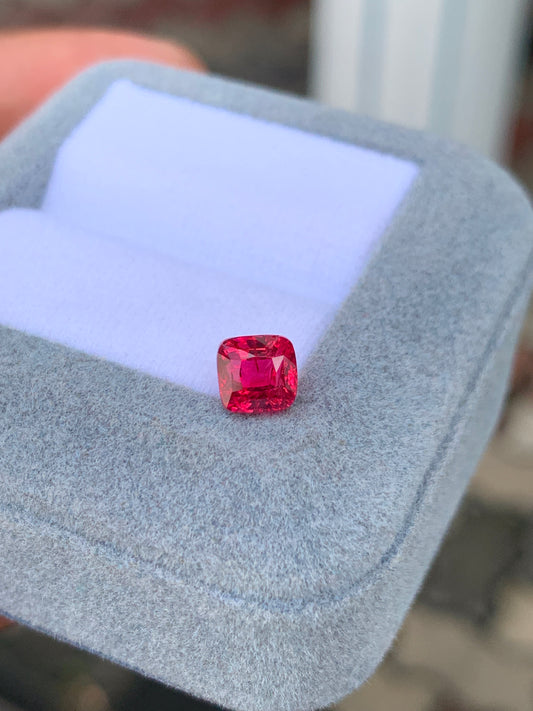 Gros plan sur le Spinelle rouge de 1,54 carat de Bonnot Paris, niché dans un écrin en velours gris. Ouvert, le coffret dévoile la pierre précieuse taille carrée sur un fond blanc doux, avec un arrière-plan flou enchanteur.