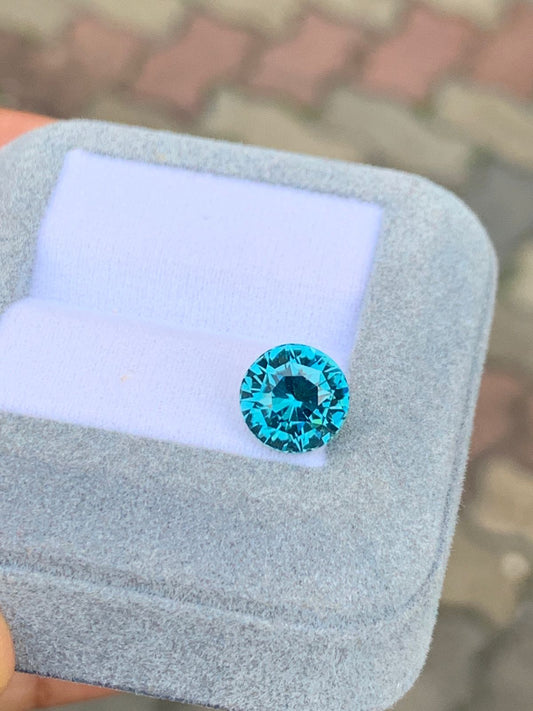 Une tourmaline bleue ronde de 3,47 carats de la maison Bonnot Paris est présentée dans un écrin de velours bleu clair ouvert. Ses facettes scintillantes se détachent sur un fond flou, évoquant les paysages enchanteurs de Madagascar.