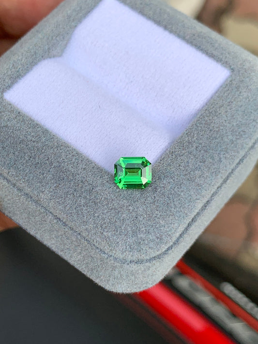 Une tsavorite vibrante Bonnot Paris (1,07 ct) de taille émeraude repose sur une boîte à bijoux grise avec un intérieur blanc, sur un fond flou.