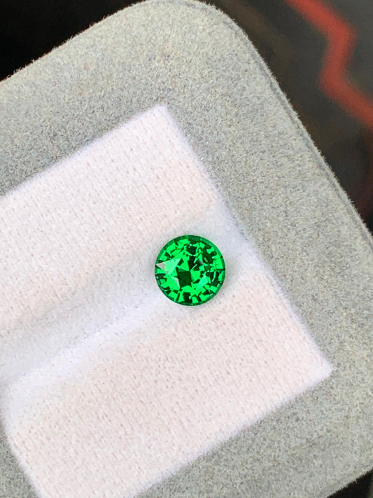 Une tsavorite vert émeraude éclatante de 1,60 carat, parfaitement ronde et facettée, est présentée sur une surface blanche matelassée dans un écrin gris. Provenant de Tanzanie et provenant de [NOUVEAU] Bonnot Paris, elle scintille sous la lumière de ses reflets verts profonds.