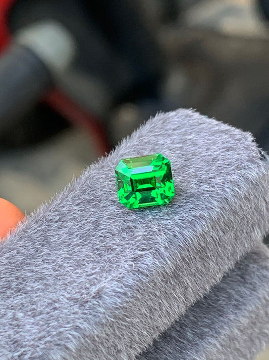 Gros plan sur une tsavorite de 2,01 carats de [NOUVEAU] Bonnot Paris, une pierre précieuse, posée sur un tissu gris doux. L'émeraude dévoile sa couleur verte éclatante et sa taille facettée.