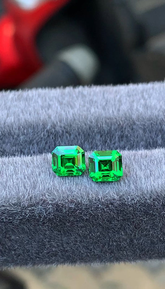 Deux tsavorites taille émeraude éclatantes de 2,27 ct, signées [NOUVEAU] Bonnot Paris, sont placées côte à côte sur un doux tissu gris. Originaires de Tanzanie, elles scintillent de clarté et dévoilent des facettes complexes et une couleur intense.