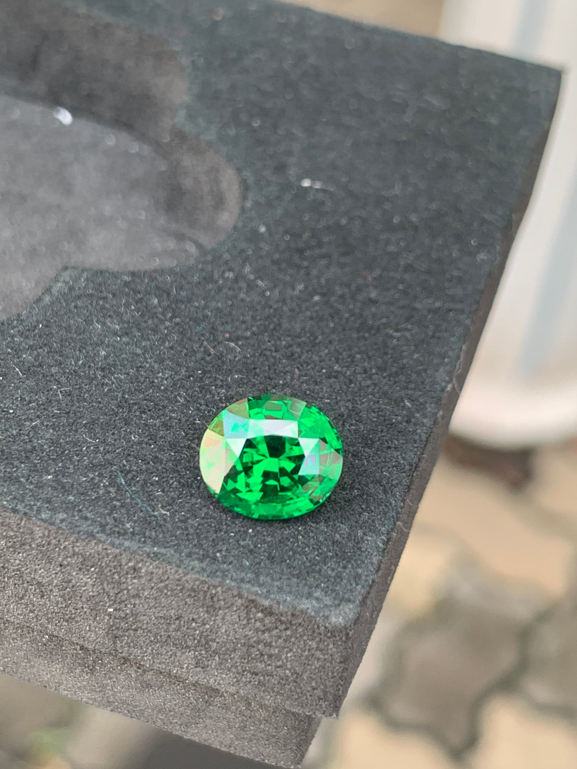 Une Tsavorite de 3,10 carats de [NOUVEAU] Bonnot Paris, provenant de Tanzanie, repose sur une surface noire texturée, sa forme ovale reflétant brillamment la lumière de ses facettes complexes.
