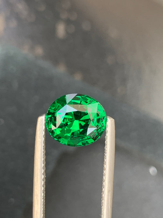 Gros plan sur une tsavorite de 3,10 carats de Bonnot Paris, tenue par une pince en argent. Cette gemme d'un vert vif scintille sous la lumière, révélant ses facettes expertes, sa couleur captivante et sa clarté sur un fond gris flou.