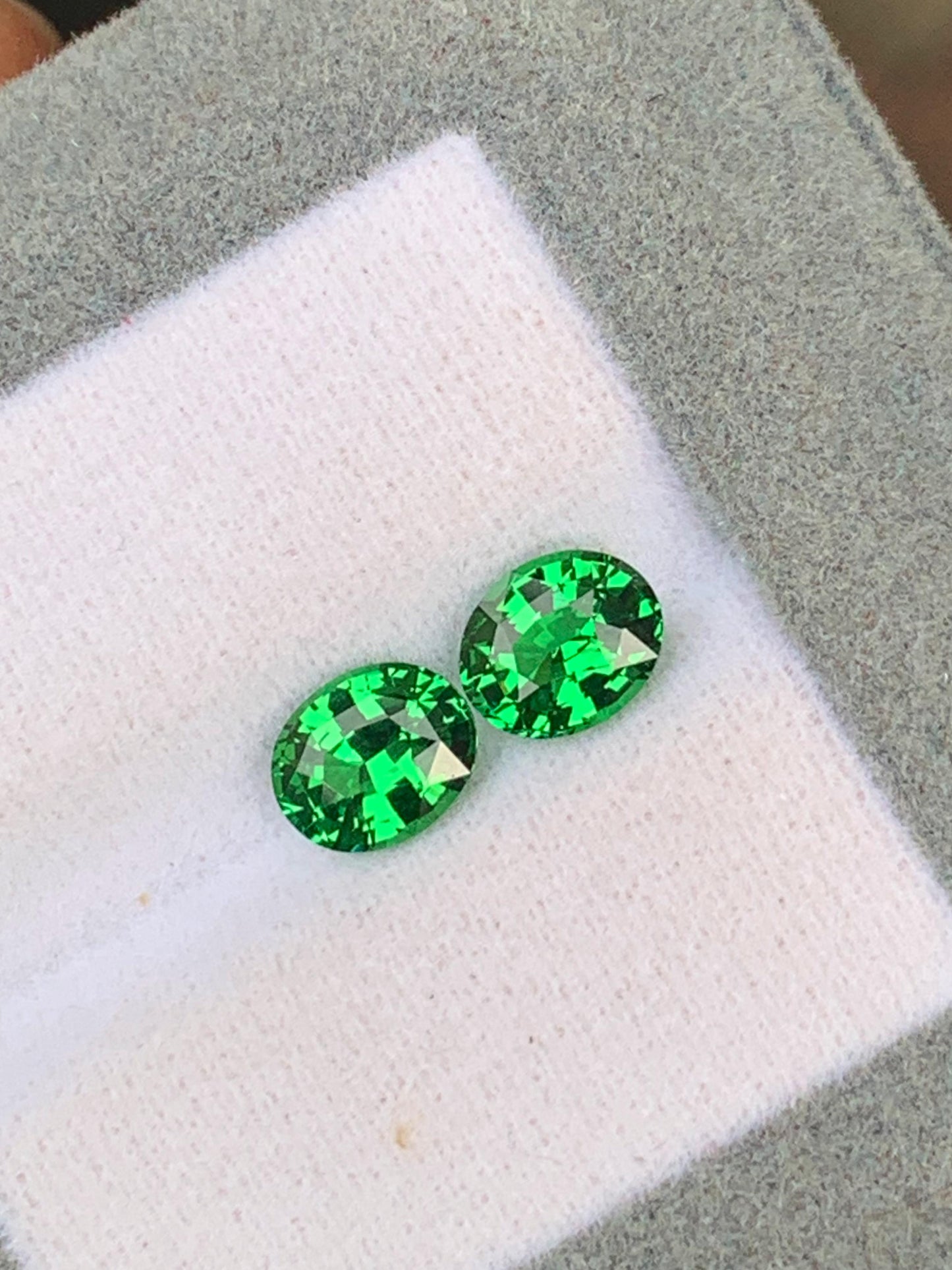Deux pierres précieuses Tsavorite taille ovale de 2,15 ct de [NOUVEAU] Bonnot Paris, connues pour leur teinte vibrante et leur brillance facettée, reposent sur un tissu blanc à l'intérieur d'une boîte de présentation doublée de gris.