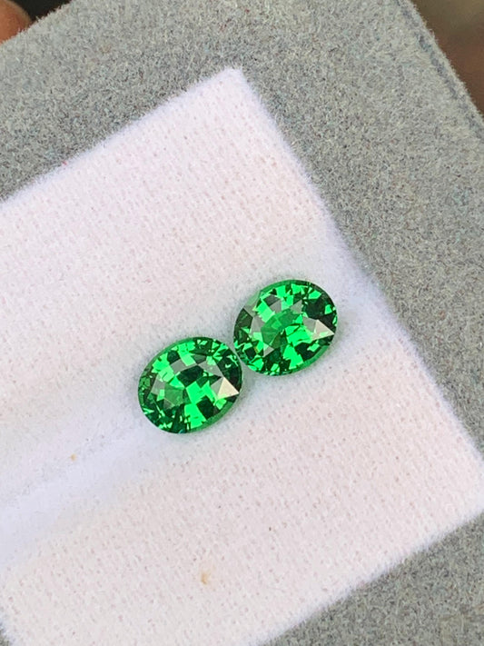 Deux pierres précieuses Tsavorite taille ovale de 2,15 ct de [NOUVEAU] Bonnot Paris, connues pour leur teinte vibrante et leur brillance facettée, reposent sur un tissu blanc à l'intérieur d'une boîte de présentation doublée de gris.