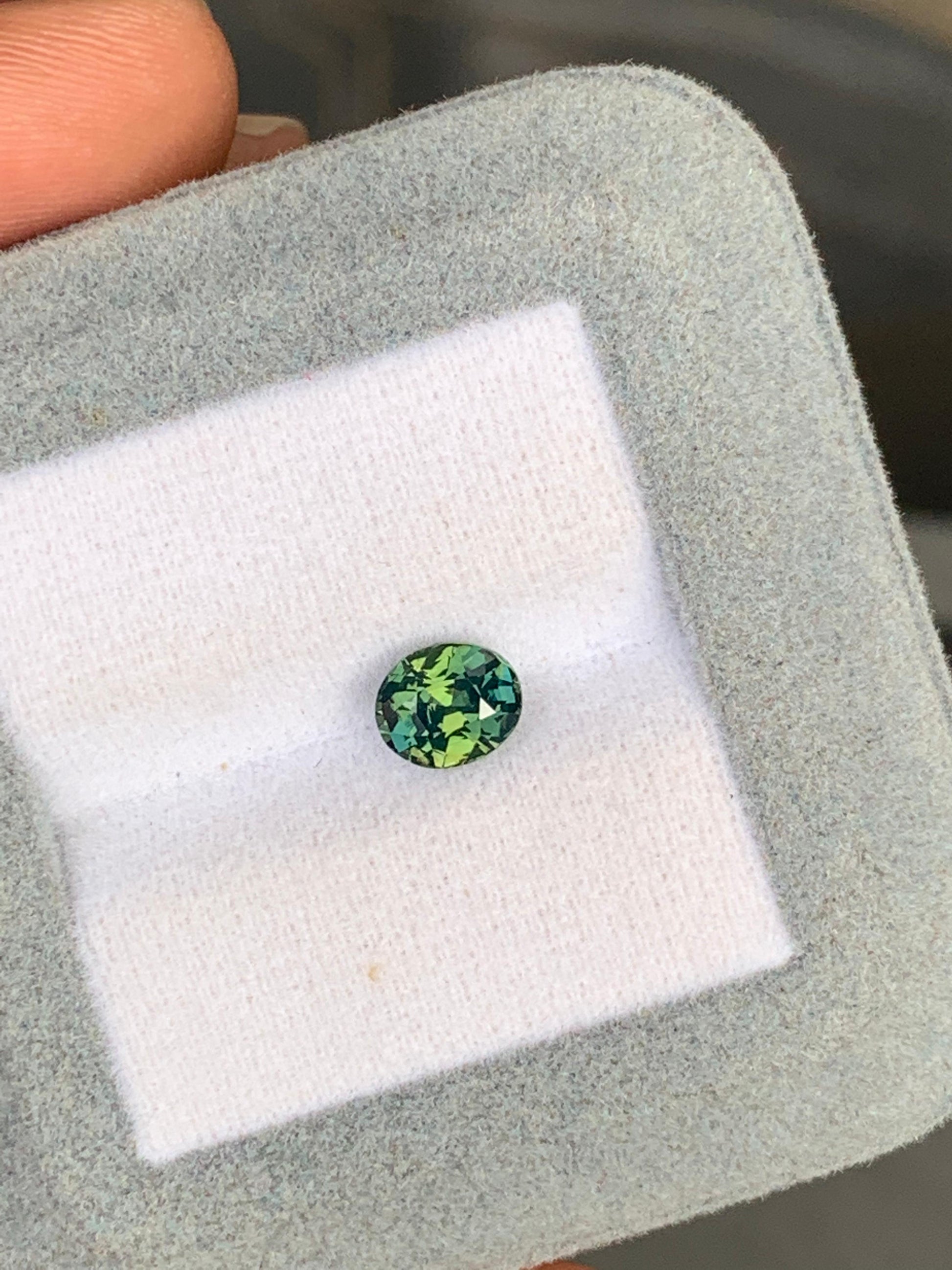 Dans un coffret à bijoux gris clair doublé de velours de chez [NOUVEAU] Bonnot Paris, un petit saphir bicolore rond de 1,51 carat scintille de nuances vertes et bleues. Ce joyau exquis du Sri Lanka est délicatement maintenu et scintille sur la surface blanche.