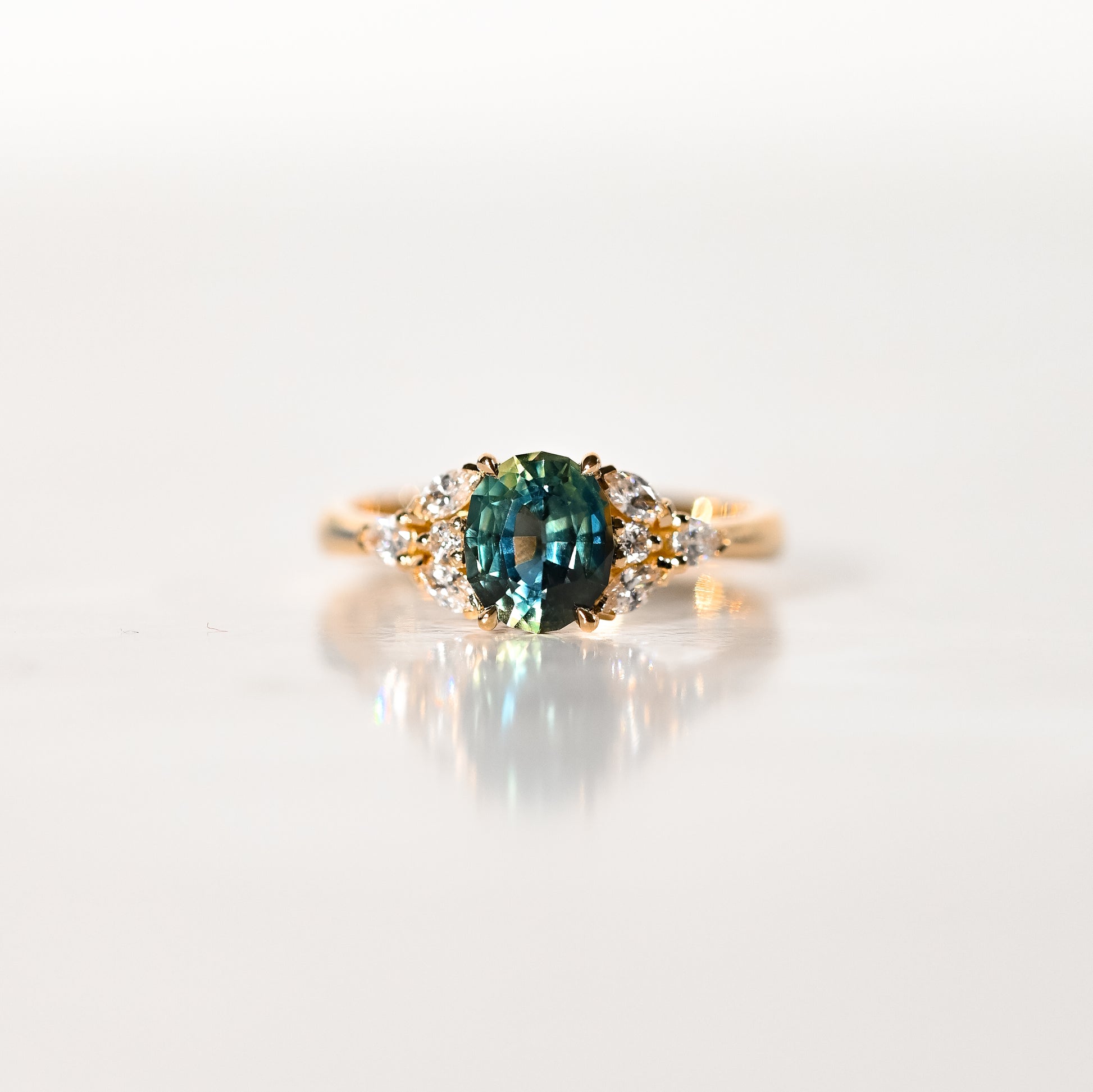 La Bague Saphir Teal · 1,5 carat de Bonnot Paris [NOUVEAU] est ornée d'un saphir ovale turquoise saisissant, encadré de diamants blancs ronds. Elle repose élégamment sur une surface blanche lisse et réfléchissante, telle une bague de fiançailles raffinée.